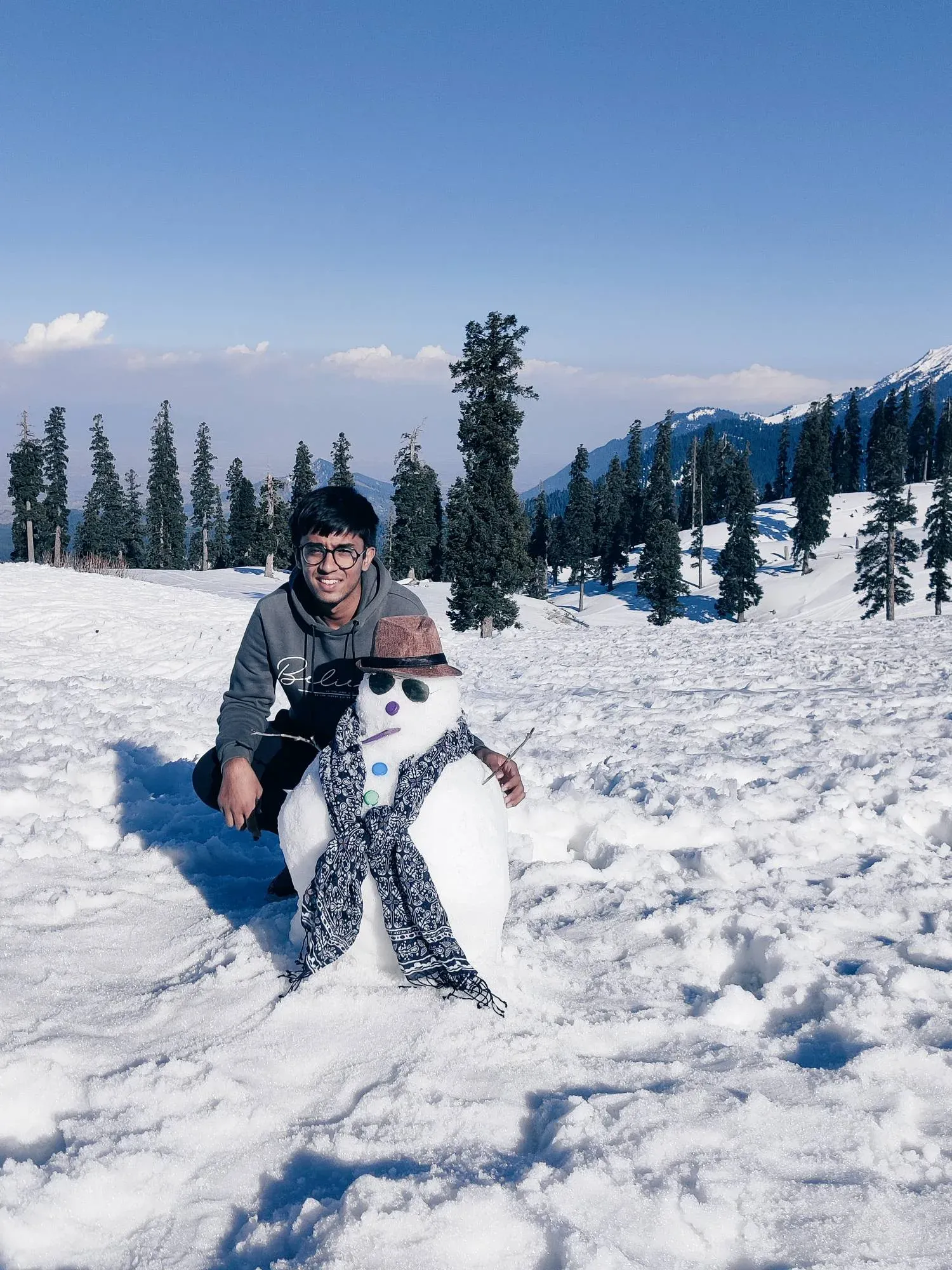Crouching beside a snowman in a hat, sunglasses and scarf